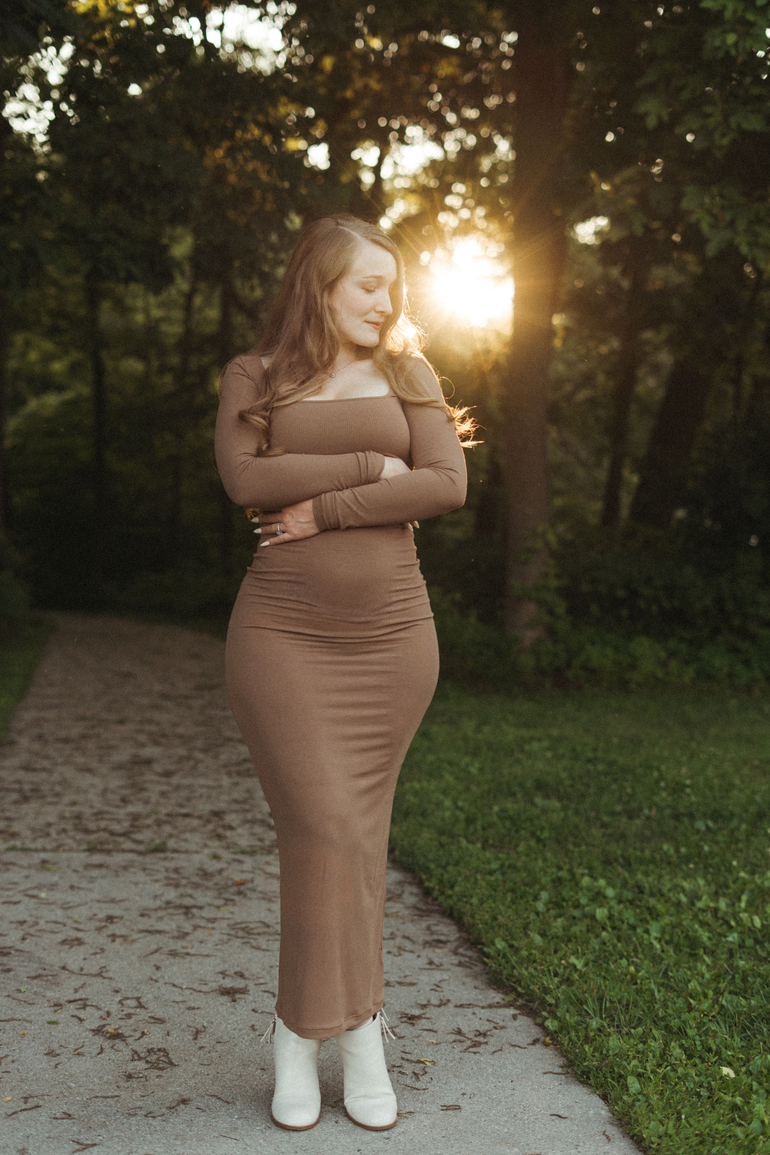 Pregnant woman in form-fitting brown off-shoulder dress poses on garden path at golden sunset.