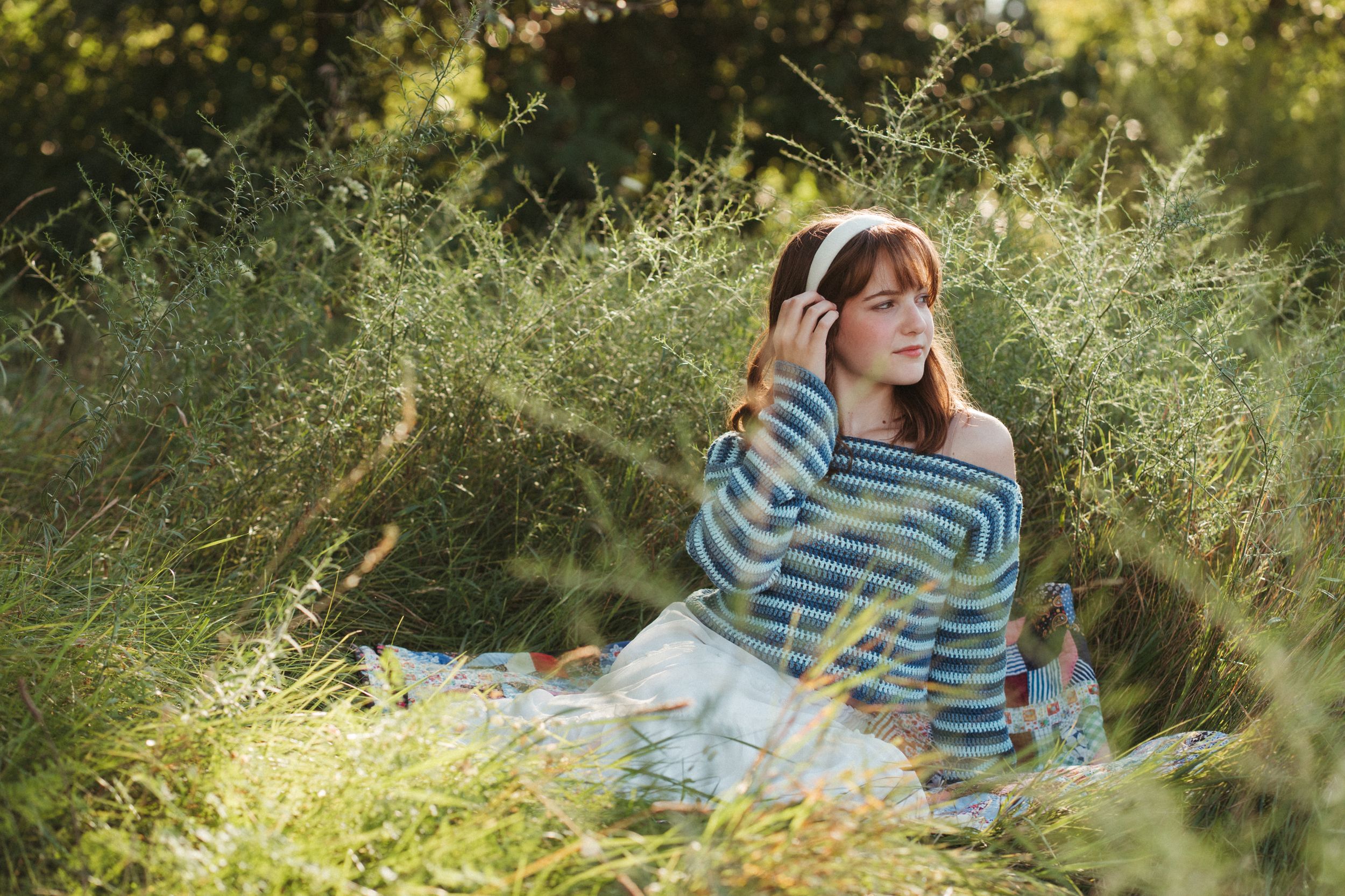 Young woman in striped off-shoulder sweater sits peacefully in lush green grass wearing white headband.
