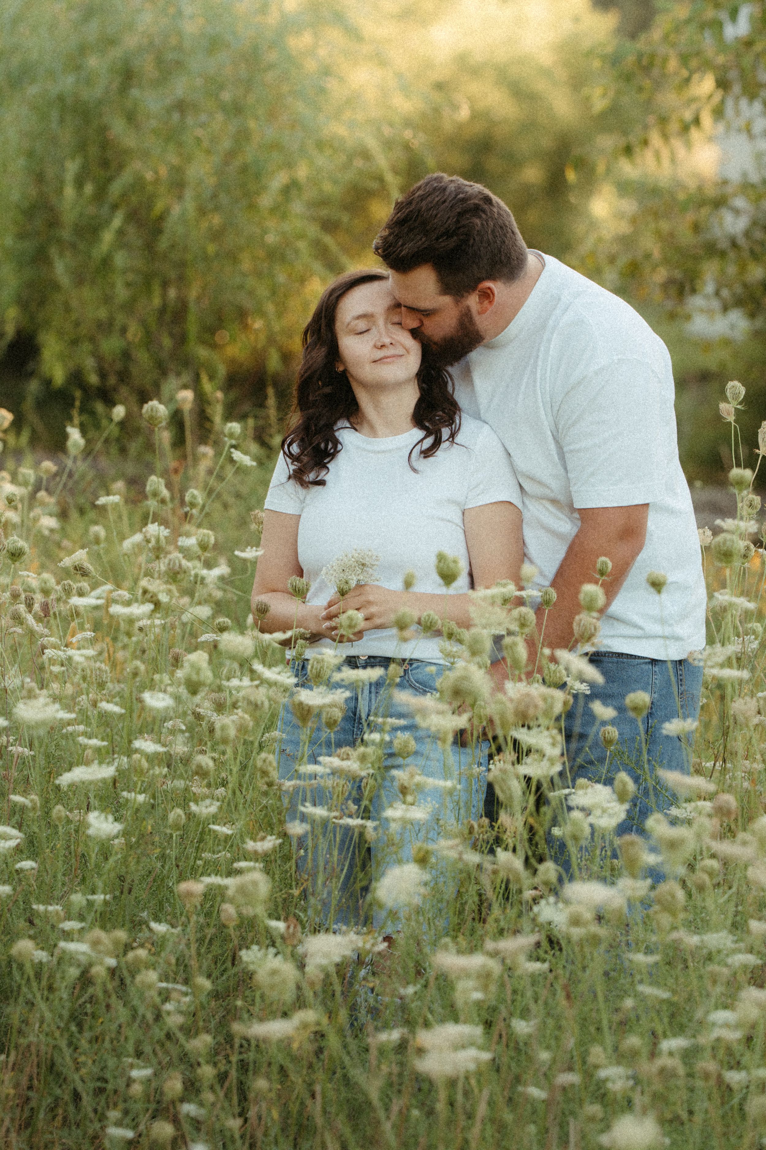 Romantic couple embracing in wildflower meadow at golden hour, man kissing womans temple tenderly.