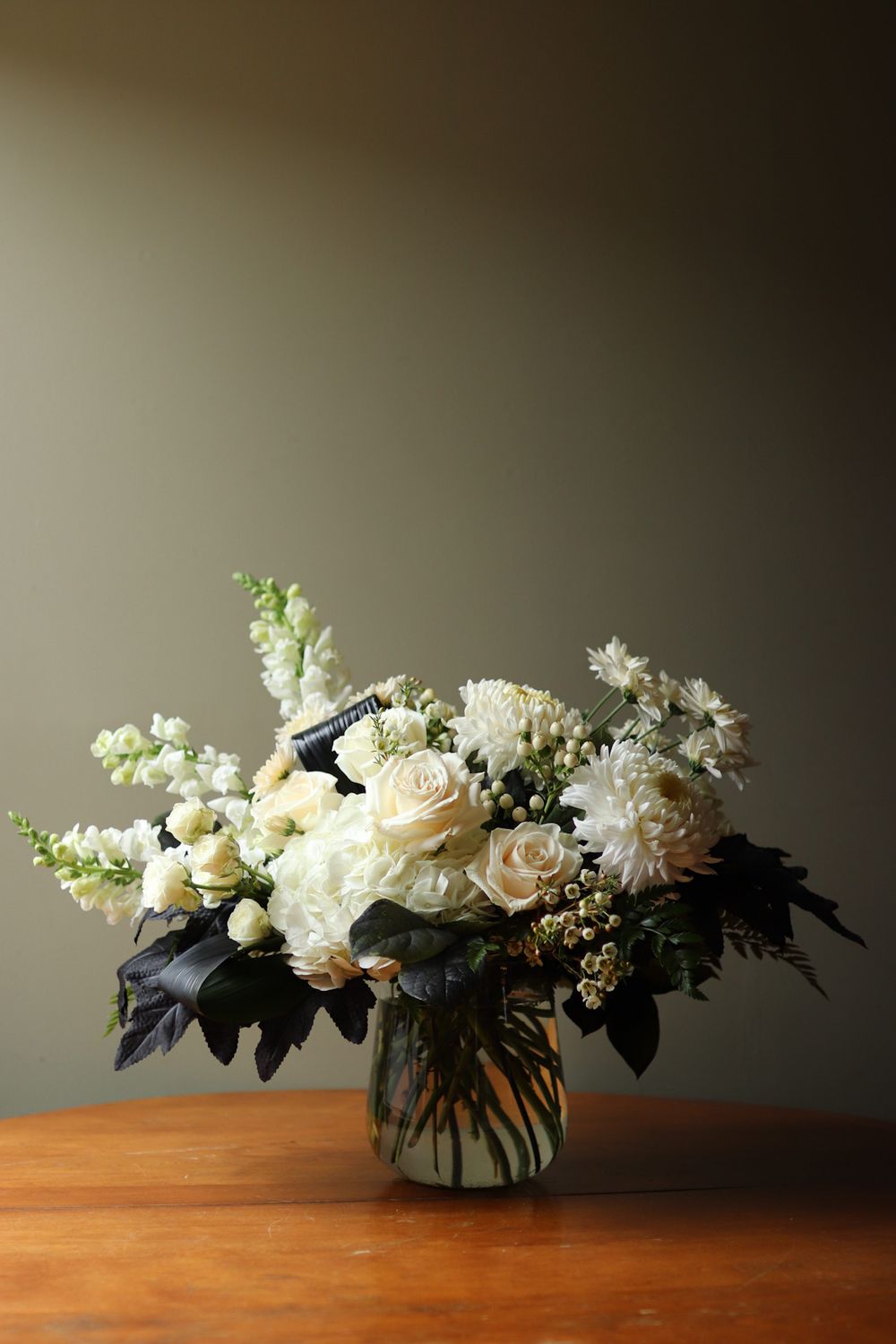 Long white floral arrangement with roses and greenery displayed on wooden table.