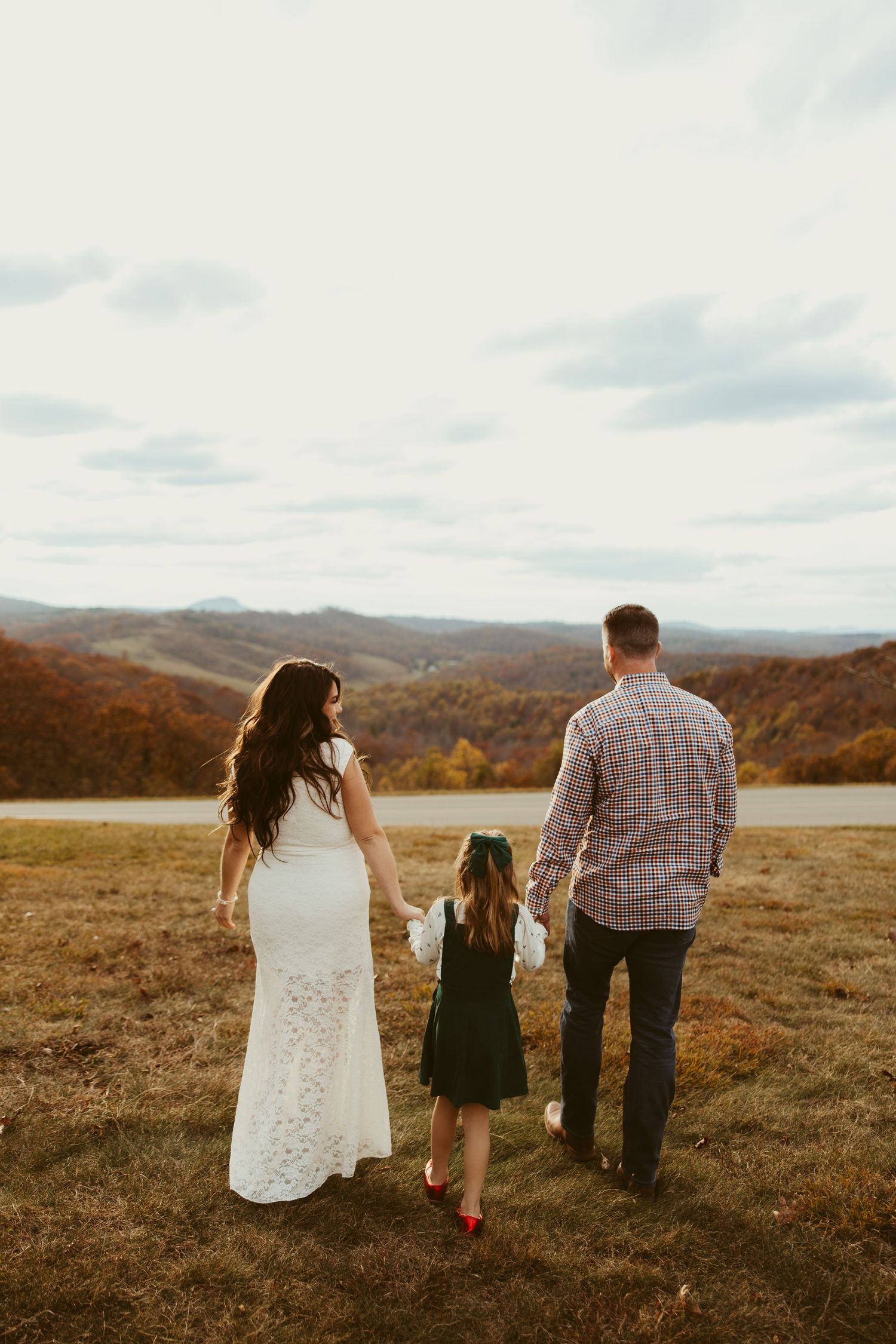 Family of three holding hands while gazing at stunning autumn mountain landscape from open field.