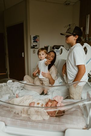 Happy family with toddler and father admiring newborn baby girl in hospital bassinet after birth.