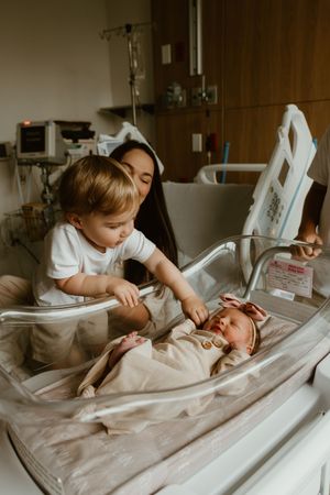 Curious toddler gently touching newborn baby sister lying in hospital bassinet with mother watching.