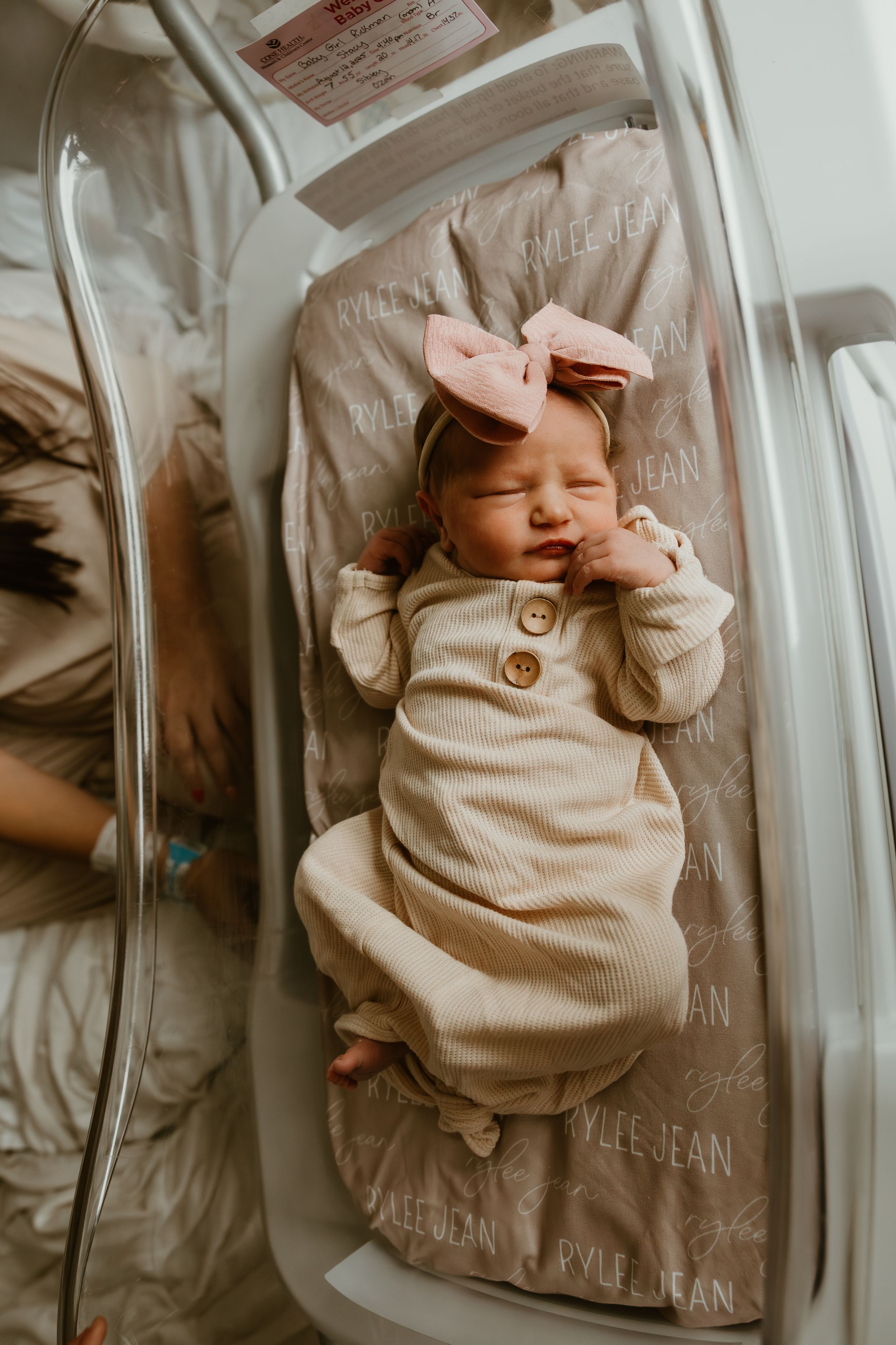 Sleeping newborn baby girl in beige romper and pink bow on personalized name bassinet sheet.