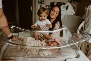 Proud mother and smiling toddler sibling looking at newborn baby in clear hospital bassinet.