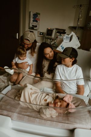 Family gathered around newborn baby in hospital bassinet, parents and siblings sharing a tender first meeting moment.
