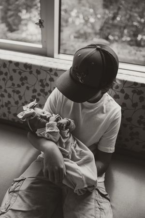 Father in baseball cap tenderly holding newborn baby in black and white hospital photo.