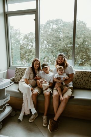 Happy family of five posing by hospital window with newborn baby, siblings smiling together.