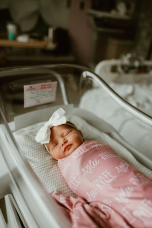 Sleeping newborn baby girl swaddled in pink personalized blanket with white bow headband.