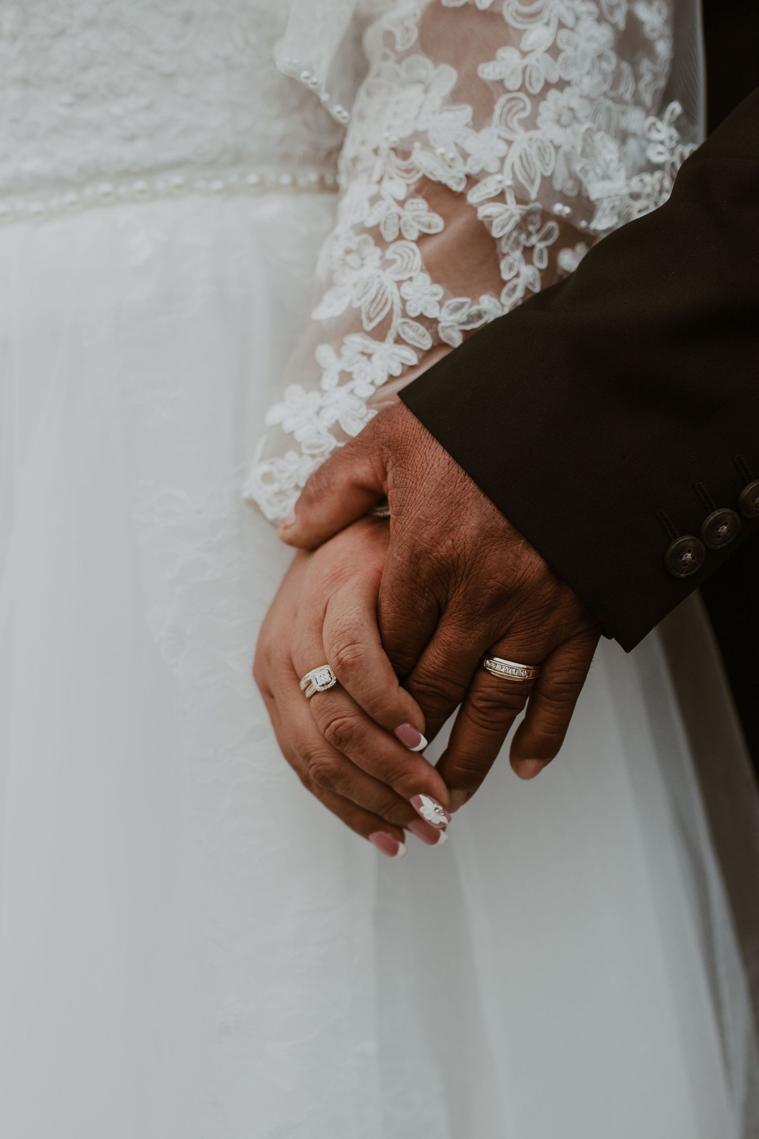 Close-up of newlyweds holding hands showing wedding rings, bride in lace gown and groom in dark suit.