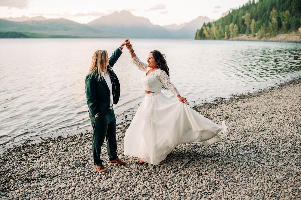 Couple in wedding attire walking along scenic mountain lake shore at Apgar Picnic Area Shoreline on pebble beach during sunset ceremony.