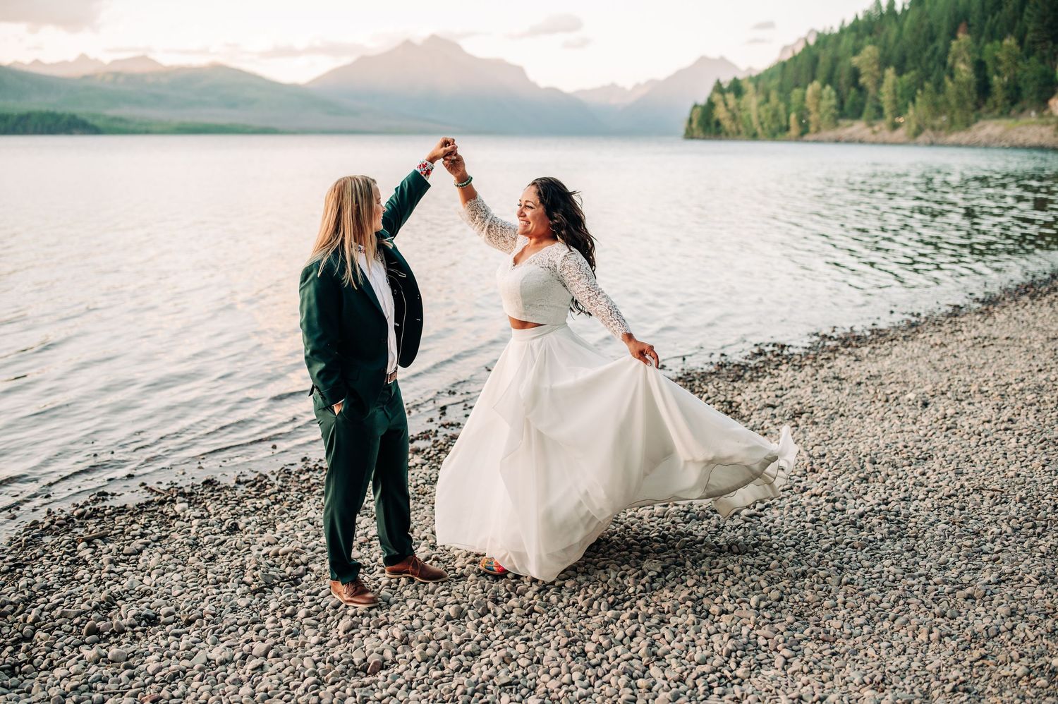 Couple in wedding attire walking along scenic mountain lake shore at Apgar Picnic Area Shoreline on pebble beach during sunset ceremony.