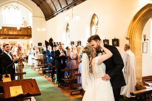 Newlywed couple shares first kiss walking down church aisle as guests applaud their wedding.
