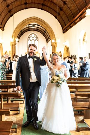 Joyful newlyweds raise hands triumphantly while walking down church aisle after wedding ceremony.