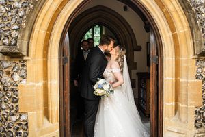 Bride and groom share romantic kiss framed by stone arched doorway of English church.
