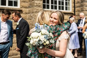 Bride in lace gown smiles while embracing a guest in a floral dress outside a stone building.