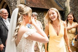 Bride in lace gown laughs with bridesmaid in yellow dress outside a sunny church after ceremony.