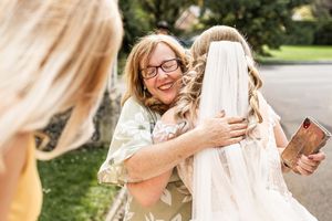 Smiling woman with glasses warmly hugs bride in white veil and gown outdoors on wedding day.