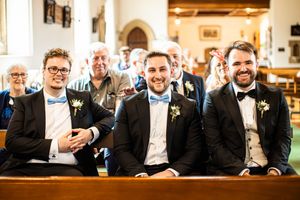 Three groomsmen in black tuxedos with blue bow ties smiling during wedding ceremony in church.