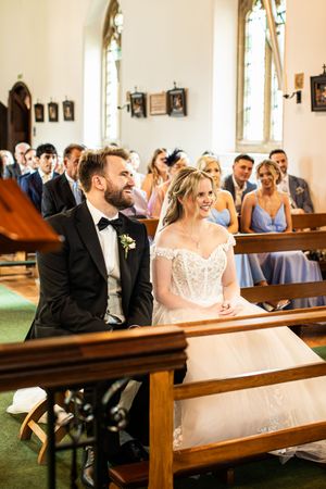 Bride and groom sit together smiling during church wedding ceremony with guests behind them.