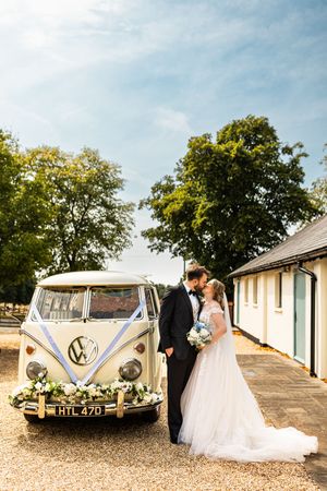Bride and groom kissing beside a classic cream VW camper van decorated with flowers outdoors.