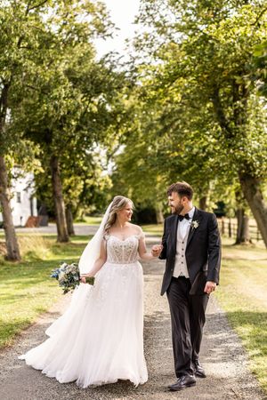 Bride and groom walking hand in hand along a tree-lined path on their sunny wedding day.
