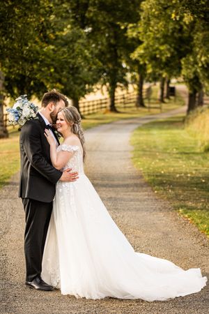 Bride and groom embracing on a scenic tree-lined gravel path on their wedding day.