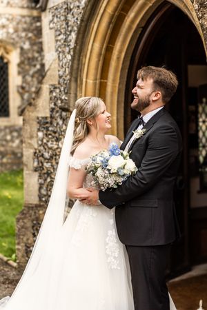 Bride and groom laugh joyfully together holding bouquet outside a stone church after their wedding.