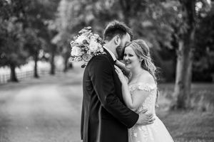 Black and white photo of bride and groom embracing on a tree-lined path with bouquet.