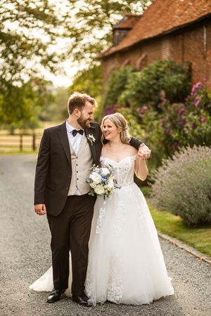 Bride and groom walking arm in arm along a gravel path beside a rustic brick building.