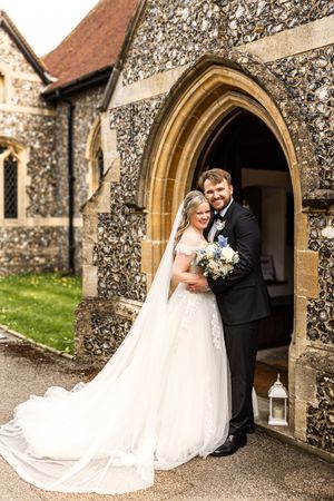 Bride and groom pose smiling under a Gothic stone church archway with long veil and bouquet.
