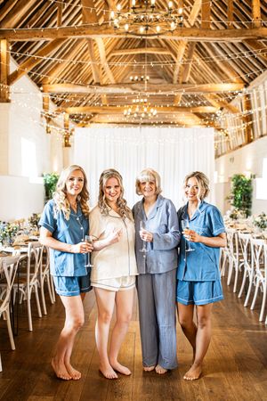 Bride and bridesmaids in matching pajamas pose with champagne inside a fairy-light decorated wedding barn.