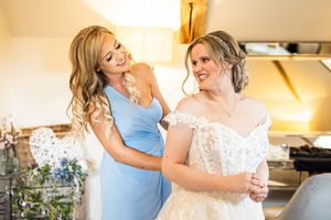 Smiling bridesmaid in blue dress helps bride into her off-shoulder lace wedding gown during preparations.