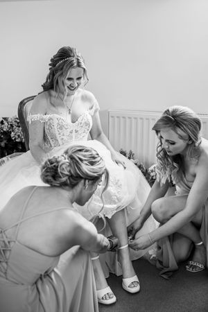 Black and white photo of two bridesmaids helping a laughing bride put on her wedding shoes.