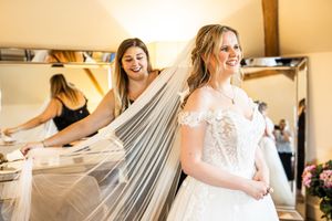 Bridesmaid spreads the veil of a smiling bride in a lace off-shoulder wedding dress during preparations.