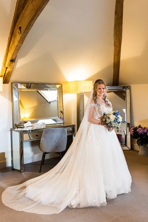 Bride in elegant lace ball gown with long veil holds bouquet in a warmly lit room with wooden beams.