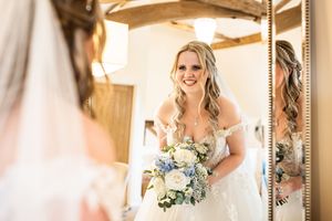 Smiling bride in off-shoulder lace gown holds blue and white bouquet while looking in a mirror.