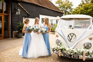 Bride and two bridesmaids in blue dresses pose beside a decorated vintage VW camper van outside.