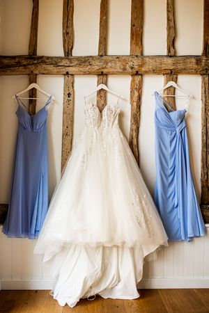 White lace wedding gown flanked by two blue bridesmaid dresses hanging on a rustic barn wall.