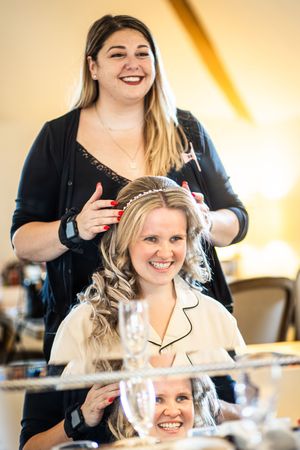 Hairstylist placing a tiara on a smiling blonde bride during wedding morning preparations.