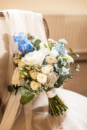 Elegant bridal bouquet of white roses and blue delphiniums resting against a chair with veil.