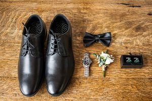 Groom accessories flat lay with black Oxford shoes, bow tie, boutonniere, watch, and cufflinks.