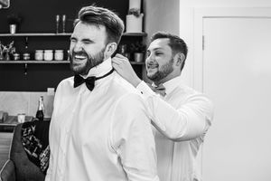 Best man helping groom adjust black bow tie in black and white wedding preparation photo.