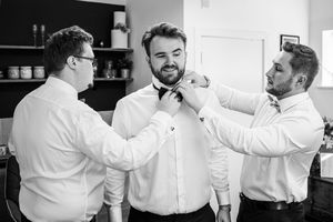 Two groomsmen helping groom fix his black bow tie during wedding morning preparations.