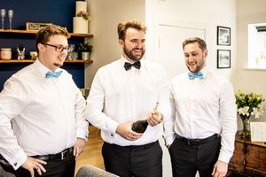 Groom and two groomsmen in blue bow ties laughing while opening champagne indoors before wedding.