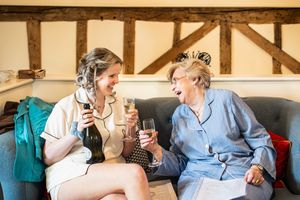Two women laughing and toasting with champagne glasses on a sofa during wedding preparations.