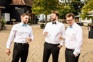 Groom in black bow tie and two groomsmen in blue bow ties laughing outdoors at wedding venue.