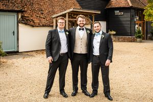 Groom in ivory waistcoat and bow tie poses with two groomsmen in black suits outside a rustic barn venue.