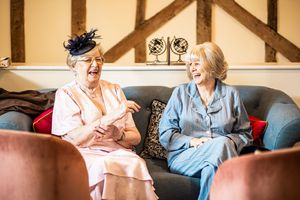 Two elderly women laugh together on a sofa in wedding guest attire inside a charming rustic venue.