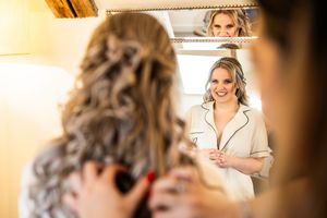 Bride in silk pajamas smiles at her reflection in a mirror during wedding morning preparations.